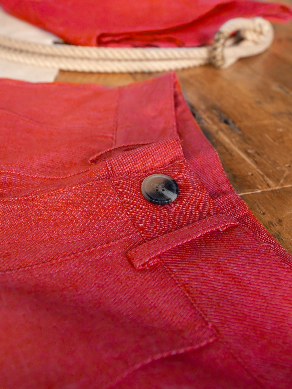 Close-up of The Tellico high waisted crop pants by Savannah Sevens western life{&}style, featuring a black button and pocket seam. The textured red fabric stands out against a rustic rope and wooden background.