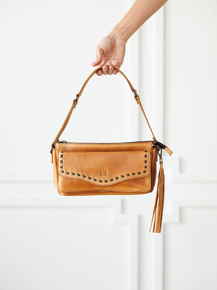 A hand holds the Temecula Leather Shoulder Bag by Savannah Sevens western life{&}style, featuring tan full-grain leather, a short strap, studded front flap, and long tassel accent against a minimalistic white background.