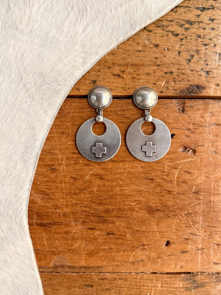 A pair of Tempe Earrings by Savannah Sevens western life{&}style, featuring sterling silver crosses with circular studs and round pendants with cut-out ovals and raised cross designs, displayed on wood next to white fur fabric.