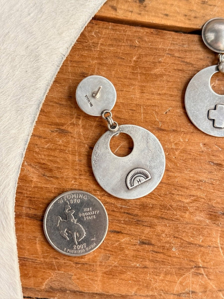 The Tempe Earrings by Savannah Sevens western life{&}style, round silver with cutout and stamped sun design, are shown next to a 1979 Wyoming quarter for scale on a wooden surface partially covered by light fur.