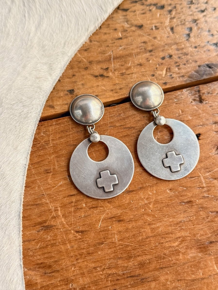 The Tempe Earrings by Savannah Sevens western life{&}style are round, handmade sterling silver cross earrings with flat disks and oval pendants, featuring a cut-out and raised cross design. Shown on wood beside white textured fabric. Peyote Bird-inspired craftsmanship.
