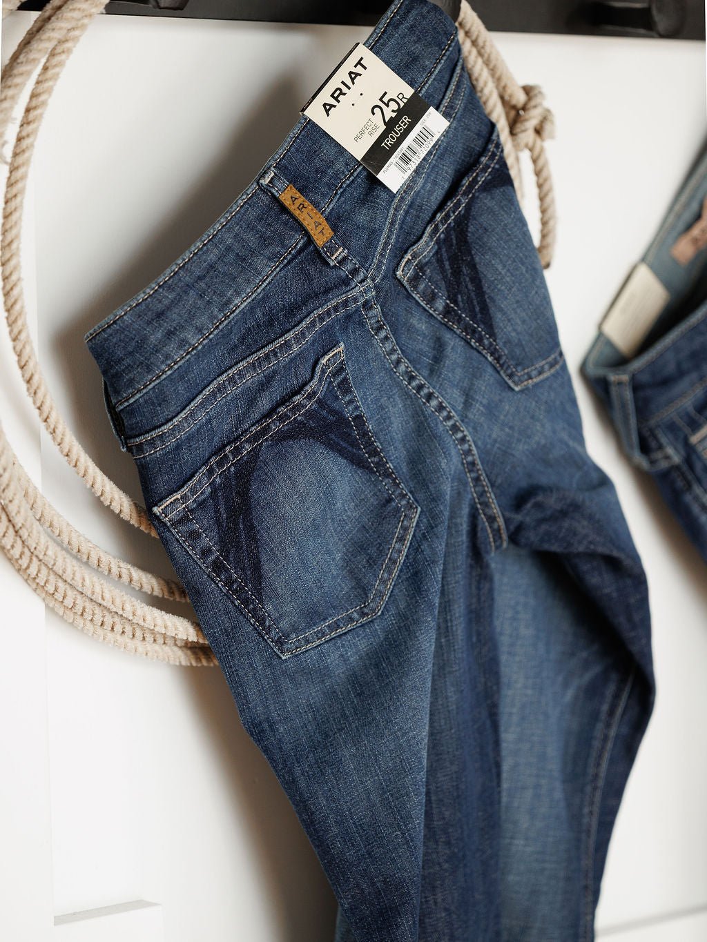 The Ariat Tex Perfect Rise Trouser Jean in medium dark wash denim hangs on a white wall beside a rope. A price tag is attached to the waistband, and the back pockets show off distinctive dark stitching.