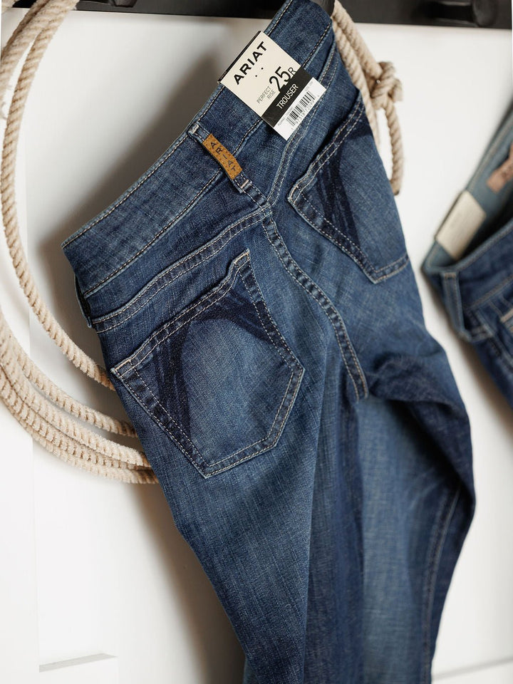 The Ariat Tex Perfect Rise Trouser Jean in medium dark wash denim hangs on a white wall beside a rope. A price tag is attached to the waistband, and the back pockets show off distinctive dark stitching.