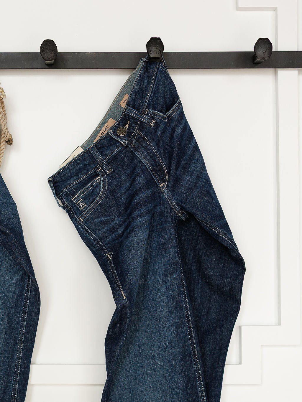 A pair of Ariat The Tex Perfect Rise Trouser Jeans in medium dark wash denim hangs by the waistband from a black hook on a white paneled wall.