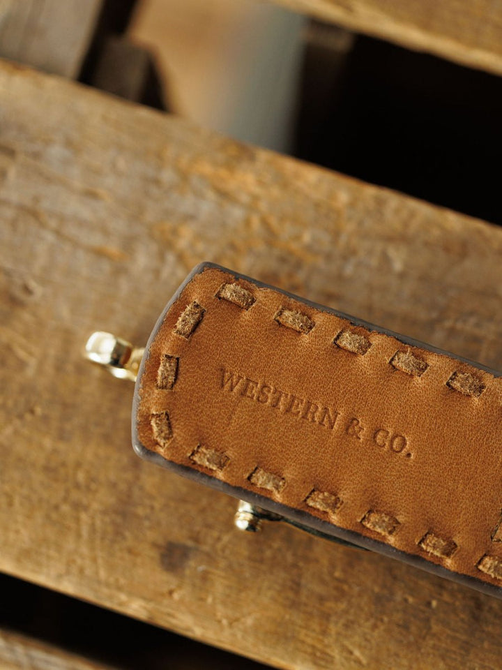 Close-up of the saddle tan Texarkana Belt by Savannah Sevens western life & style, featuring visible stitching and a stamped logo, resting on rustic wood—ideal for an authentic western look.