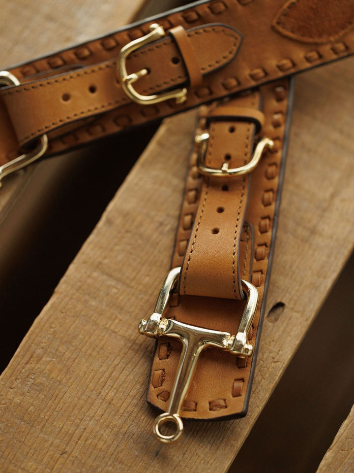 Close-up of two Texarkana Belts by Savannah Sevens western life{&}style in saddle tan leather with gold buckles, square stitching patterns, and shiny metal hardware, evoking the look of a leather snaffle bit belt on a wooden surface.