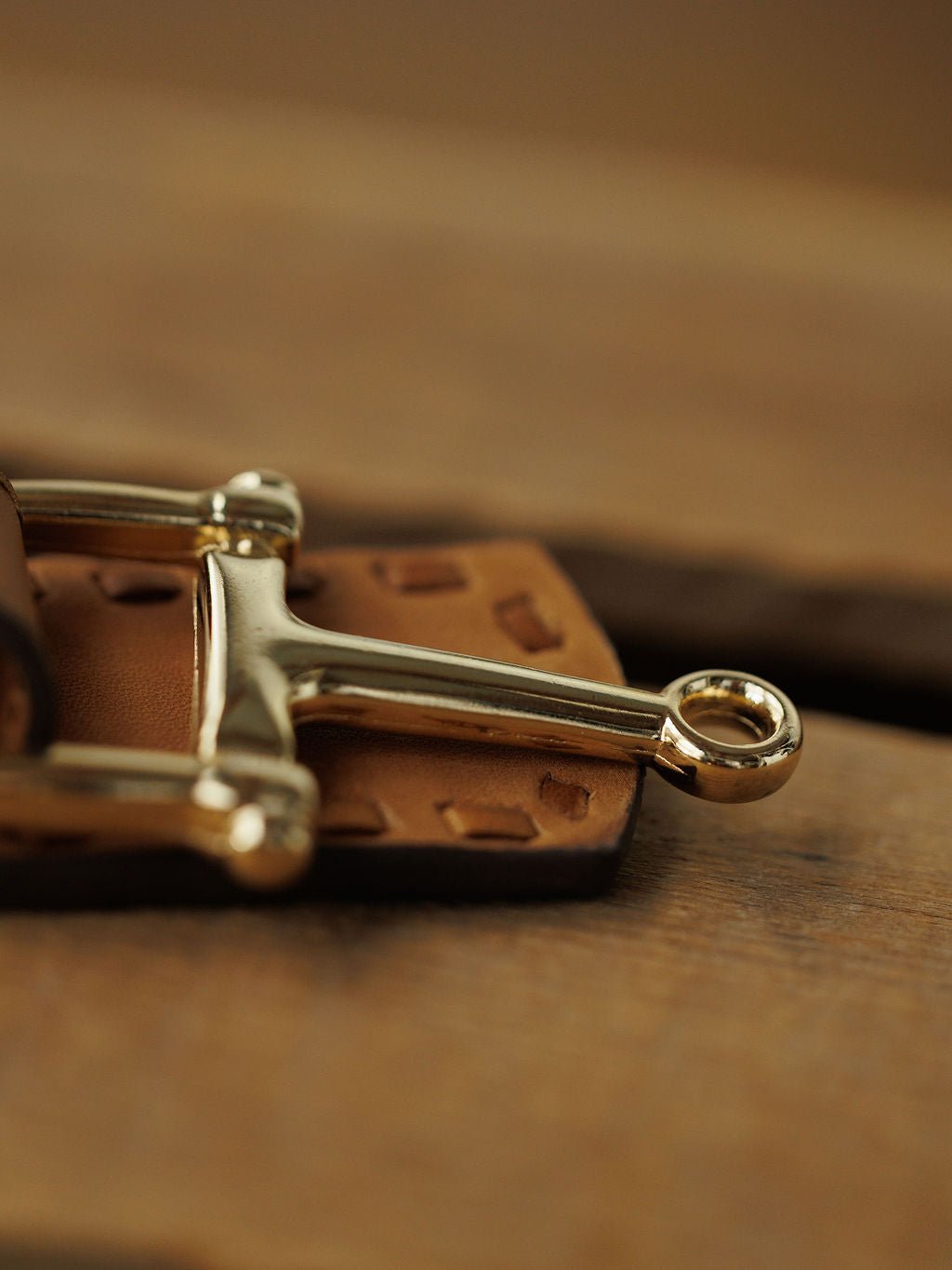 Close-up of the Texarkana Belt by Savannah Sevens western life{&}style, featuring a shiny metal horse bit on saddle tan leather with thick stitching. The blurred wood background highlights the rustic craftsmanship of this leather belt.