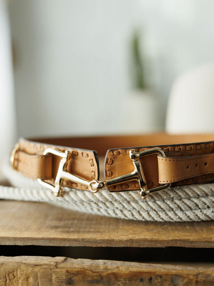 A close-up of the Texarkana Belt by Savannah Sevens western life{&}style in saddle tan leather, featuring gold buckle details, resting on a coiled white rope atop rustic wood with a blurred background.