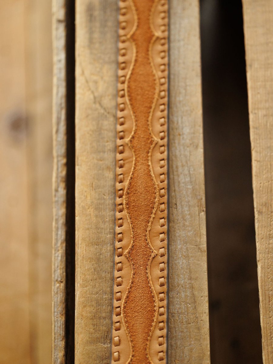 A detailed view of the Texarkana Belt by Savannah Sevens western life{&}style, featuring decorative saddle tan leather with a wavy pattern and square imprints, displayed on rustic wooden planks.