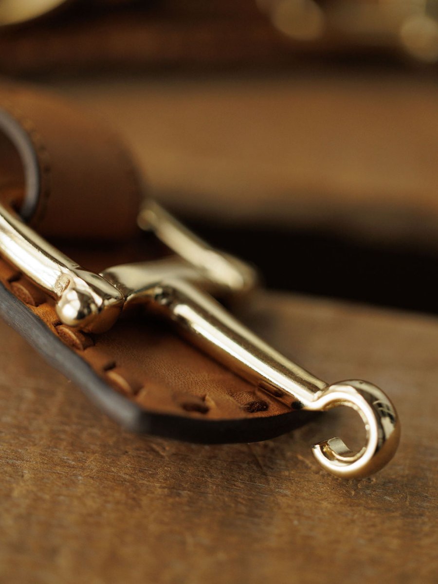 Close-up of a shiny gold metal hook on saddle tan leather with visible stitching, part of the Texarkana Belt by Savannah Sevens western life{&}style, resting on a wooden surface.