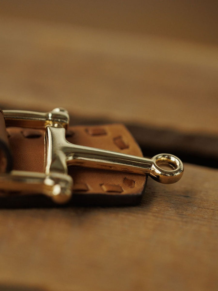 Close-up of the Texarkana Belt by Savannah Sevens western life{&}style, featuring a shiny metal horse bit on saddle tan leather with thick stitching. The blurred wood background highlights the rustic craftsmanship of this leather belt.