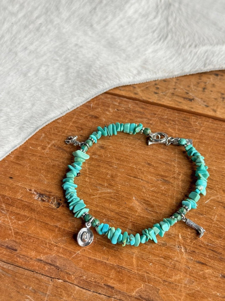 The Texas City Bracelet by Savannah Sevens western life{&}style, featuring turquoise beads and sterling silver charms, rests on a wooden surface next to light cowhide.