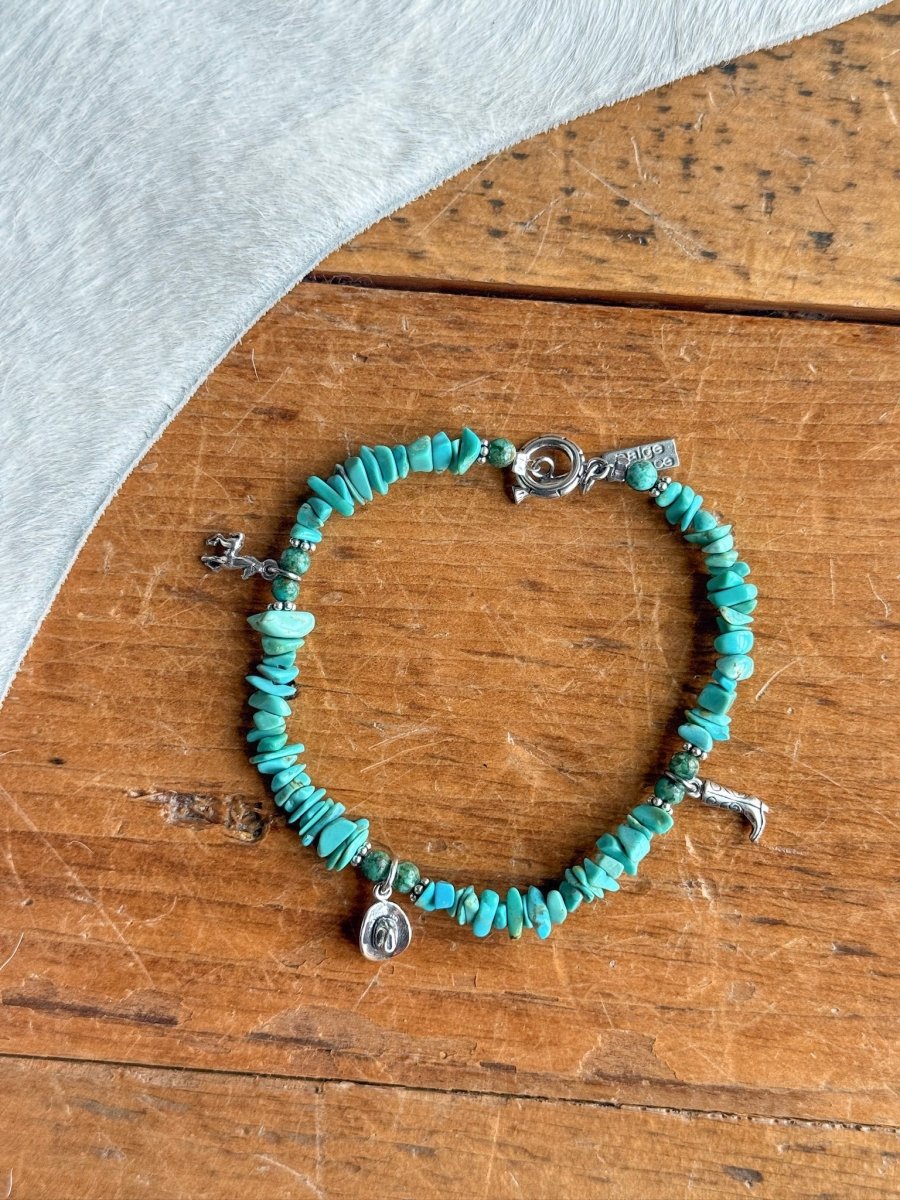 The Texas City Bracelet by Savannah Sevens western life{&}style features turquoise beads and small sterling silver charms—a boot, a cowboy hat, and a horse—displayed on wood with light gray textured fabric.