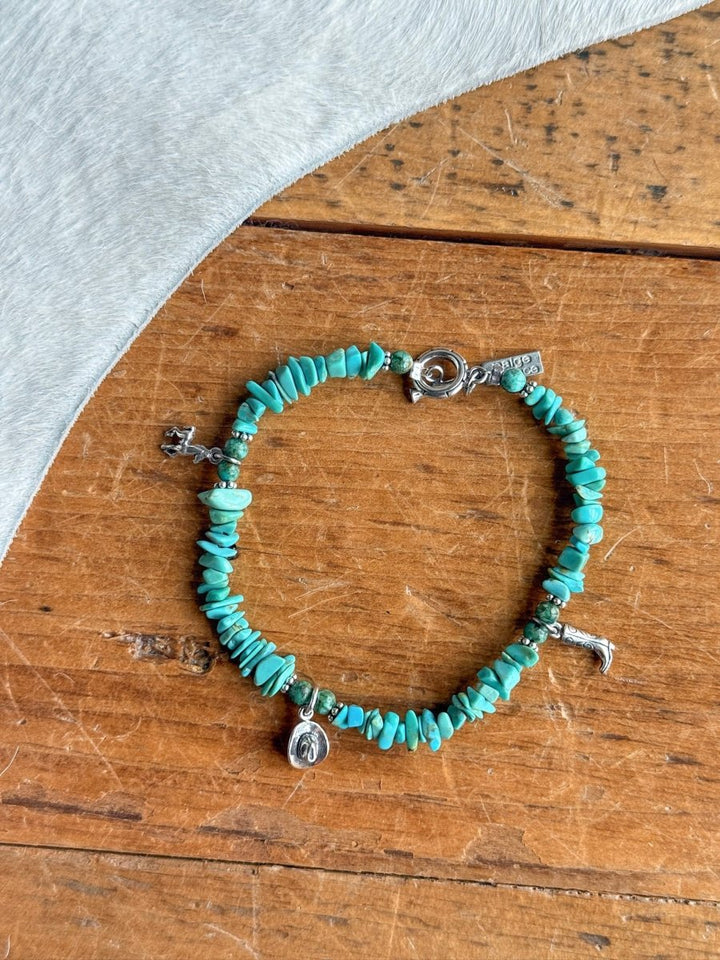 The Texas City Bracelet by Savannah Sevens western life{&}style features turquoise beads and small sterling silver charms—a boot, a cowboy hat, and a horse—displayed on wood with light gray textured fabric.