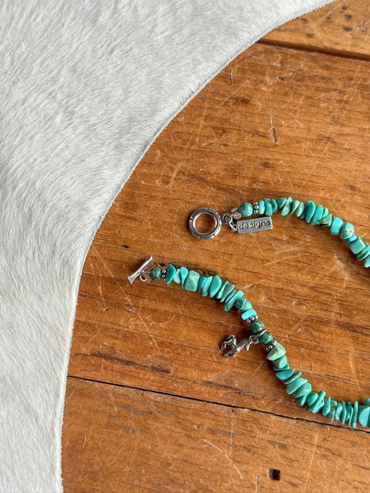 The Texas City Bracelet by Savannah Sevens western life{&}style, featuring turquoise stone beads and sterling silver charms, rests on a wooden surface near a white cowhide.