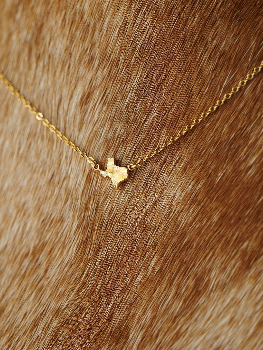 The Savannah Sevens western life{&}style Texas Necklace features a gold Texas-shaped charm pendant, resting on a brown fur-like background. Hypoallergenic for comfortable wear.