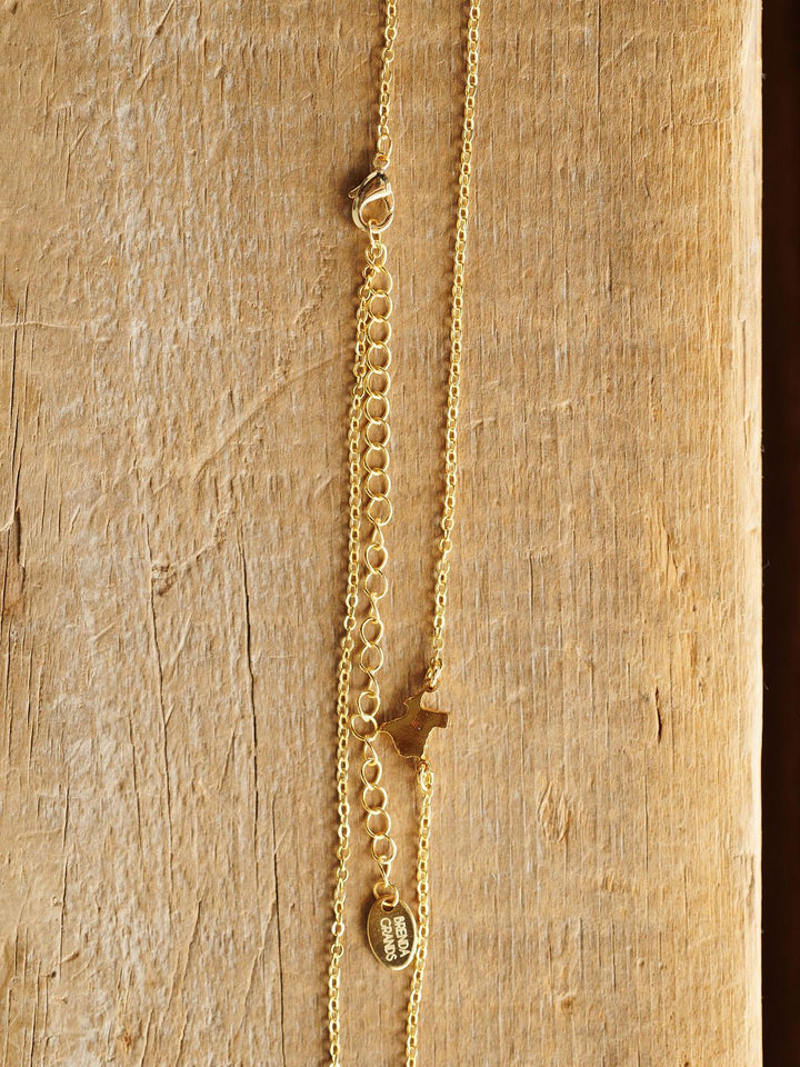 The Texas Necklace by Savannah Sevens western life{&}style features a gold Texas pendant, small dog charm, flat oval tag, and adjustable clasp on a textured wooden display. Hypoallergenic for comfortable, everyday wear.