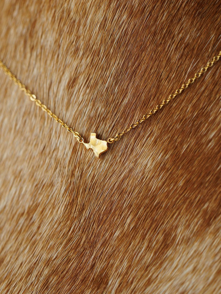 The Savannah Sevens western life{&}style Texas Necklace features a gold Texas-shaped charm pendant, resting on a brown fur-like background. Hypoallergenic for comfortable wear.