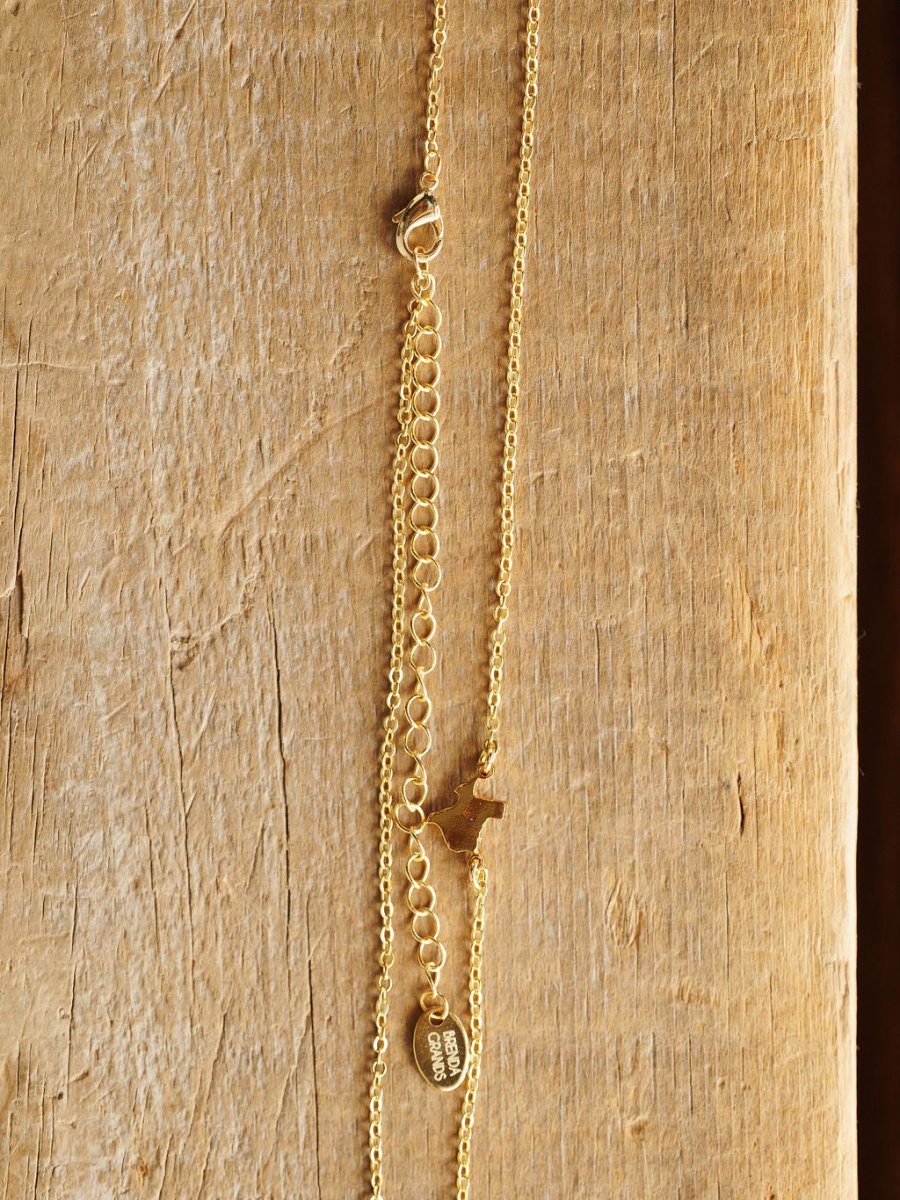 The Texas Necklace by Savannah Sevens western life{&}style features a gold Texas pendant, small dog charm, flat oval tag, and adjustable clasp on a textured wooden display. Hypoallergenic for comfortable, everyday wear.