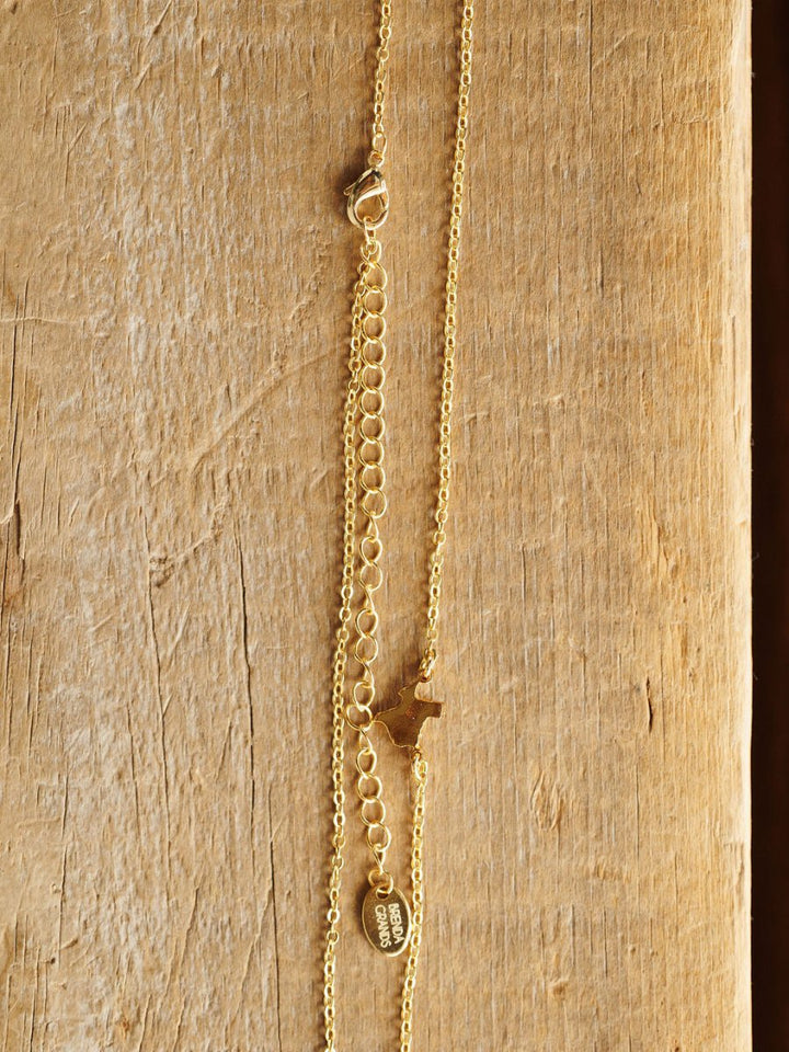 The Texas Necklace by Savannah Sevens western life{&}style features a gold Texas pendant, small dog charm, flat oval tag, and adjustable clasp on a textured wooden display. Hypoallergenic for comfortable, everyday wear.