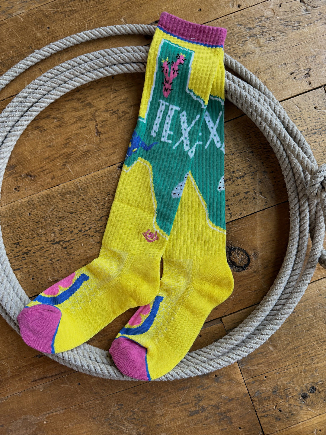 A pair of Texas Ya’ll Crew Socks by Savannah Sevens western life{&}style, featuring vibrant yellow and green colors with pink accents and patterns, are displayed inside a coiled rope on a wooden floor.