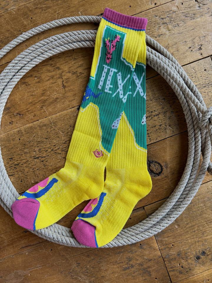 A pair of Texas Ya’ll Crew Socks by Savannah Sevens western life{&}style, featuring vibrant yellow and green colors with pink accents and patterns, are displayed inside a coiled rope on a wooden floor.