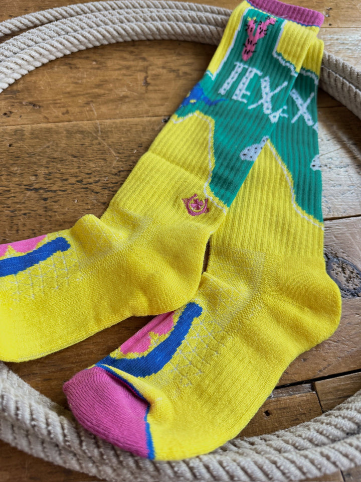 Texas Ya’ll Crew Socks by Savannah Sevens western life{&}style, featuring a colorful Texas-themed design with green, yellow, pink, and blue accents, displayed on a wooden surface within a coiled rope.