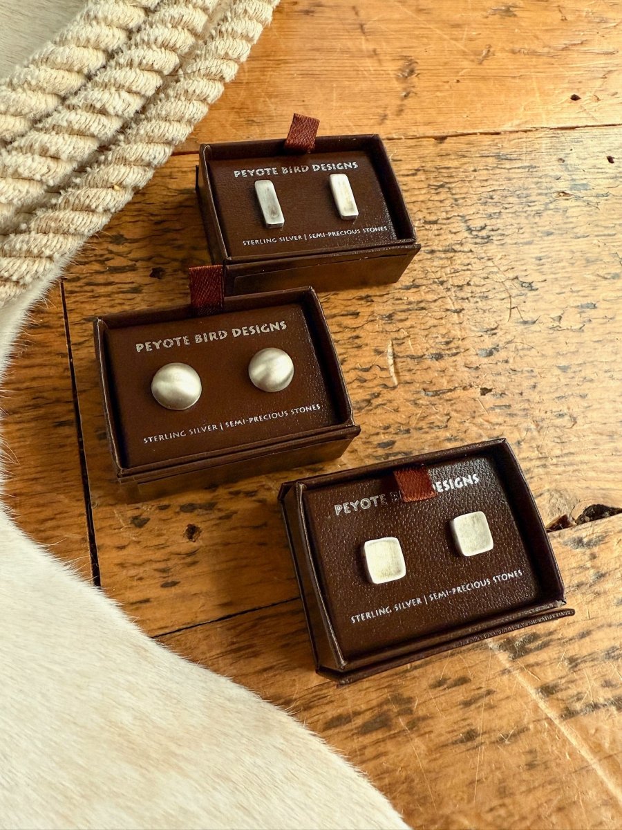 Three small brown boxes labeled Savannah Sevens western life{&}style show Texhoma Earrings—handmade sterling silver studs, one pair round and one pair square—on a wooden surface beside beige rope and white cowhide..