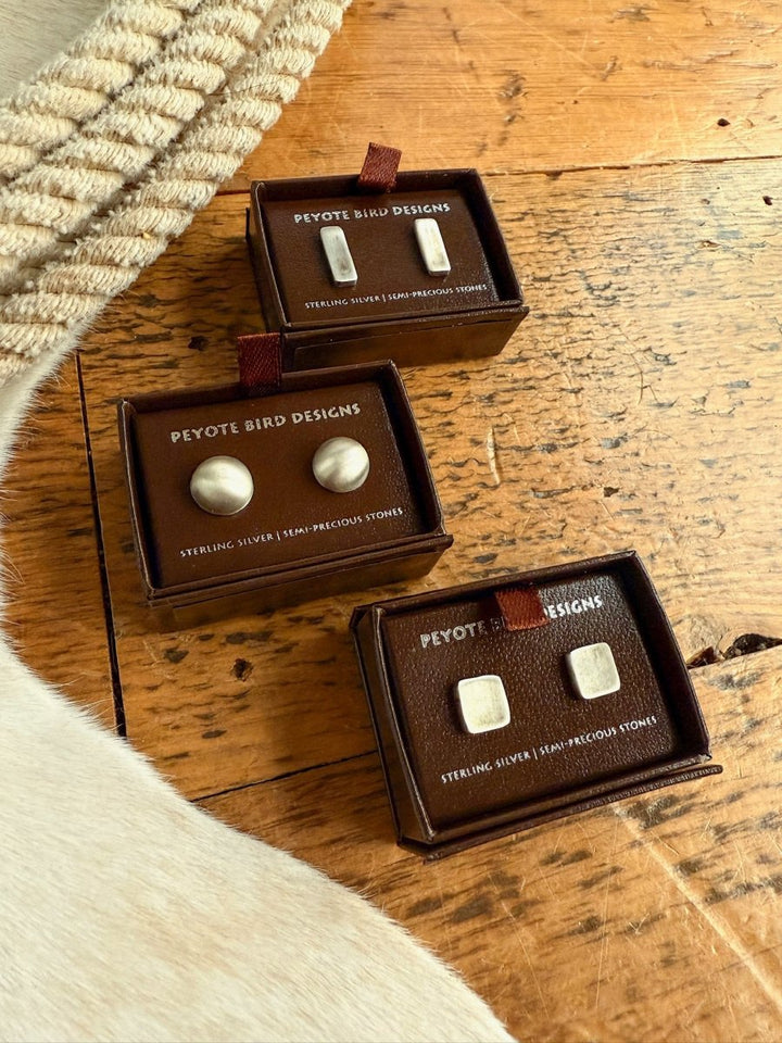 Three small brown boxes labeled Savannah Sevens western life{&}style show Texhoma Earrings—handmade sterling silver studs, one pair round and one pair square—on a wooden surface beside beige rope and white cowhide..
