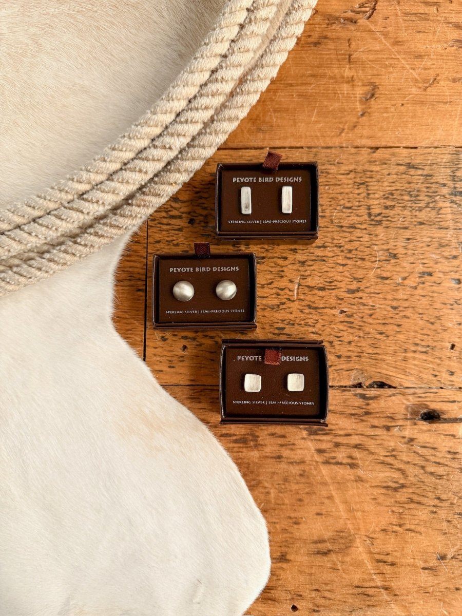 Three pairs of Texhoma Earrings by Savannah Sevens western life{&}style, including sterling silver studs, are displayed in black boxes on a wooden surface beside white textured fabric and a cream-colored fur rug.