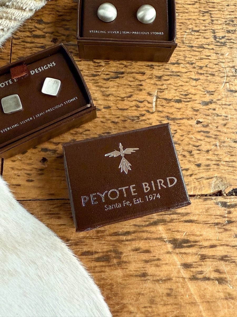 A brown Peyote Bird Designs box with a silver bird logo and “Peyote Bird Santa Fe, Est. 1974” sits on rustic wood next to two small boxes holding Savannah Sevens western life{&}style Texhoma handmade sterling silver stud earrings.