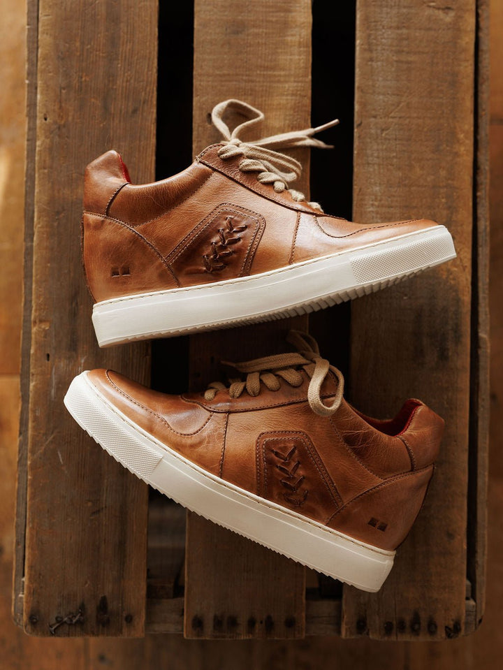 A pair of The Texicos brown leather sneakers by Savannah Sevens western life{&}style, with white soles, tan laces, decorative stitching, and a leaf-like side pattern, displayed on a rustic wooden surface. Made by BedStu. 
