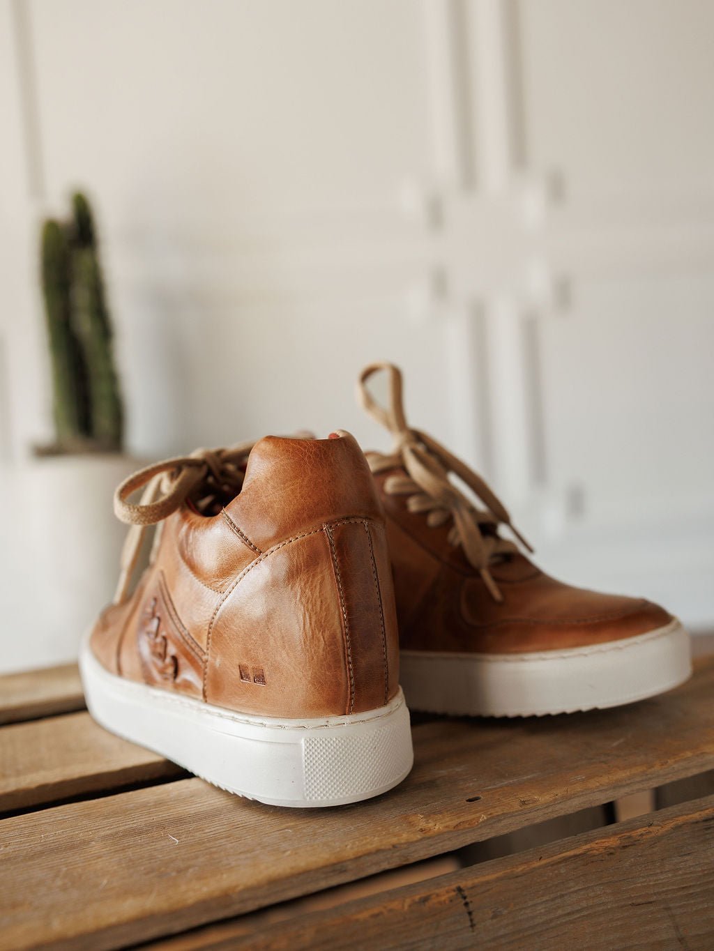 A pair of The Texicos by Savannah Sevens western life{&}style, tan leather wedge booties with white soles and beige laces, sit on a wooden surface. A blurred green cactus in a white pot appears in the background against a white wall. Made by BedStu. 
