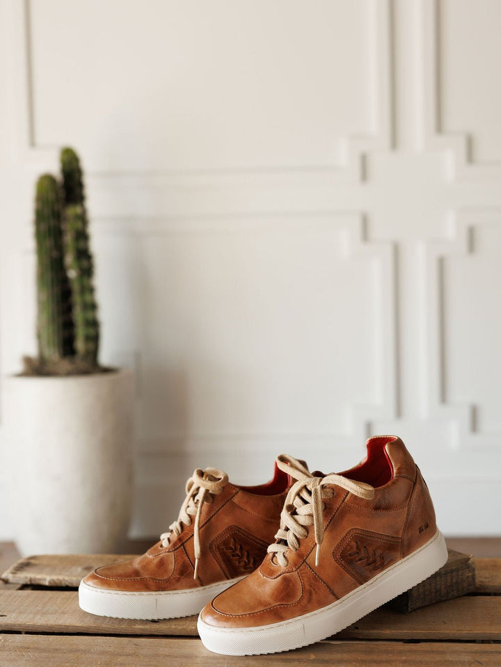 The Texicos by Savannah Sevens western life{&}style—tan leather sneakers with white soles and beige laces—are displayed on a wooden surface, with a potted cactus in the background against a white paneled wall. Made by BedStu. 