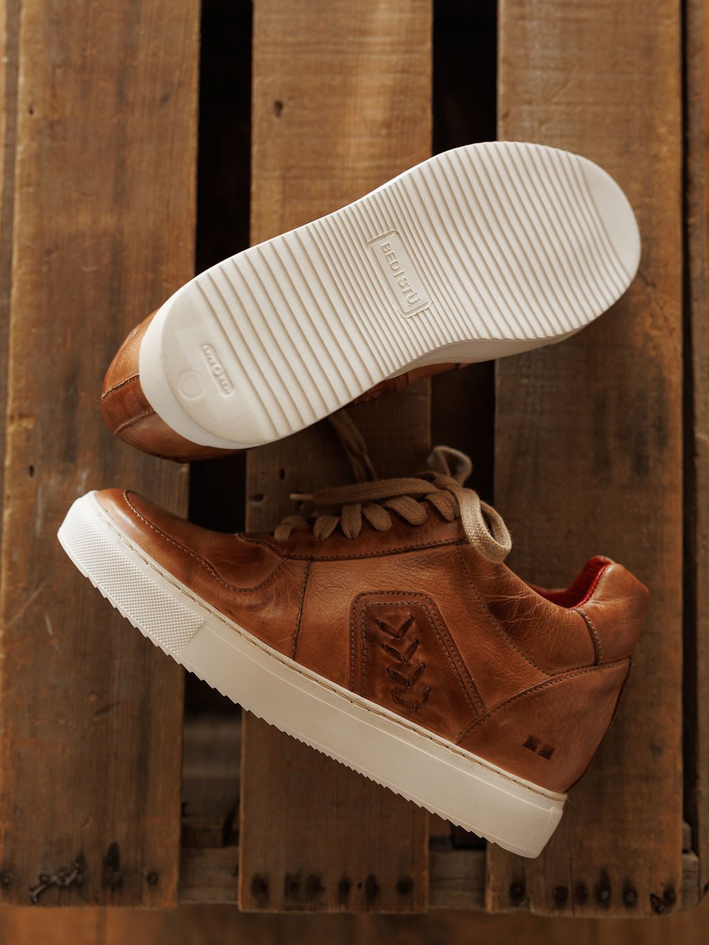 A pair of The Texicos by Savannah Sevens western life{&}style brown leather sneakers with white soles are shown on a wooden surface; one displays the tread while the other stands upright highlighting its side and laces. Made by BedStu. 