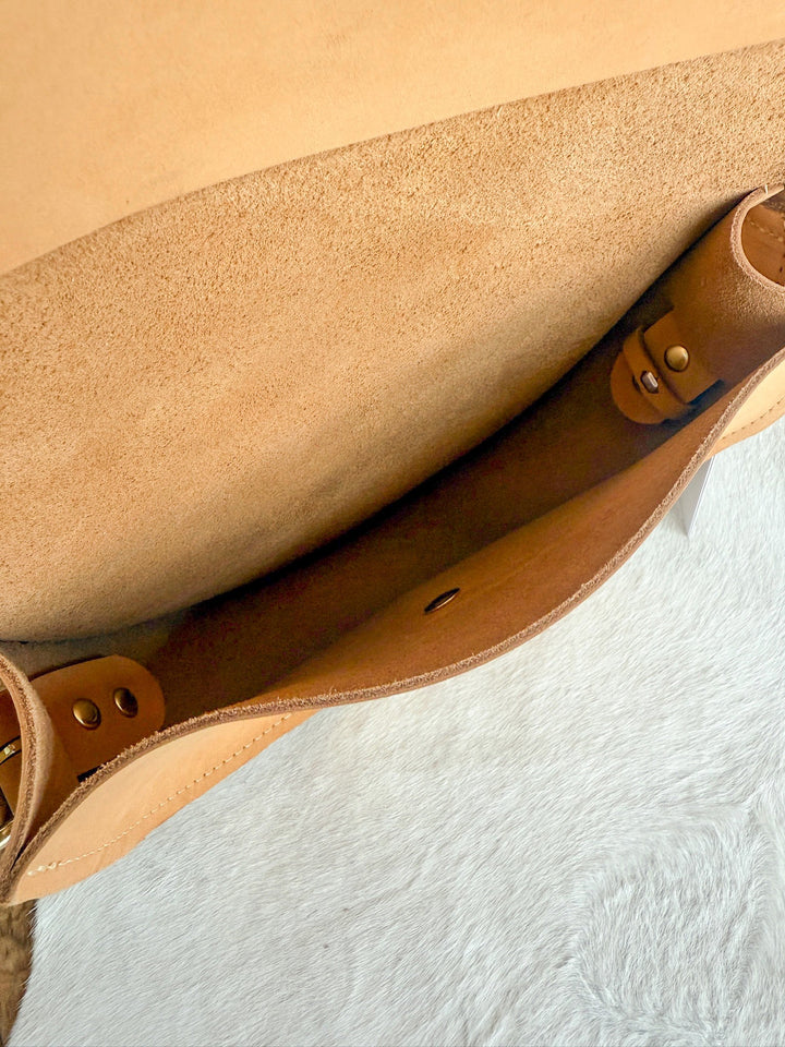 Close-up of The Texola by Savannah Sevens western life{&}style, featuring a saddle tan full-grain leather exterior, suede interior, deep open compartment, and brass rivets. Bag rests on a lightly textured surface.