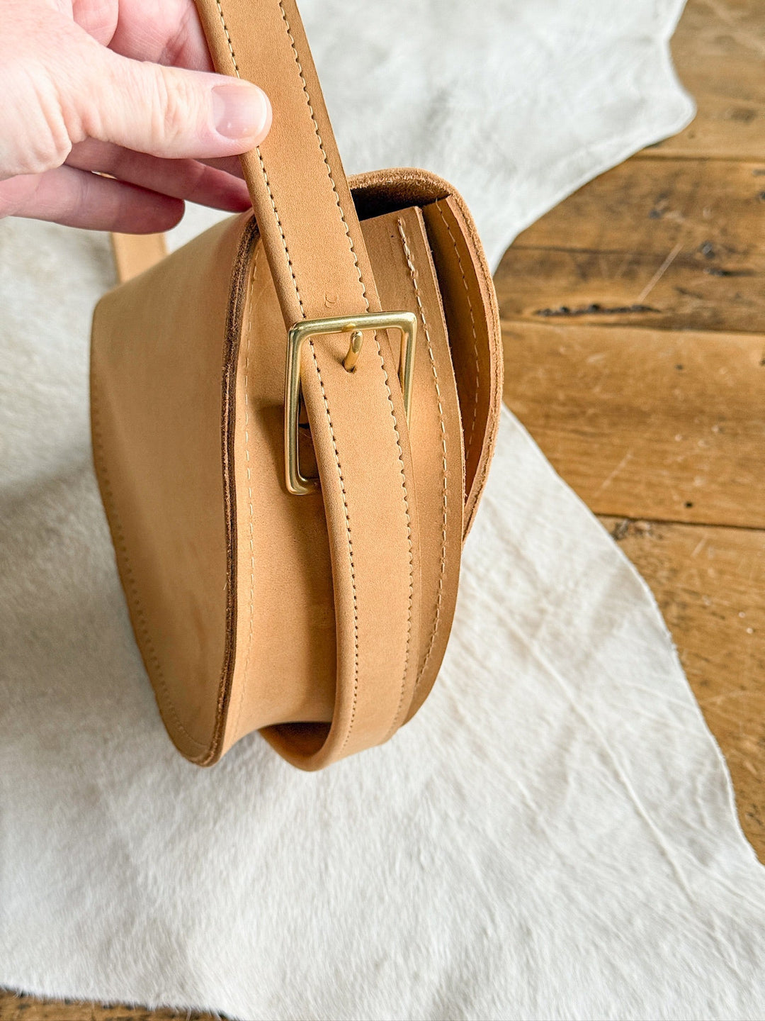 A hand holds the strap of The Texola by Savannah Sevens western life{&}style, a saddle tan full-grain leather bag with visible stitching and a gold buckle, set on light fabric on a wooden floor.