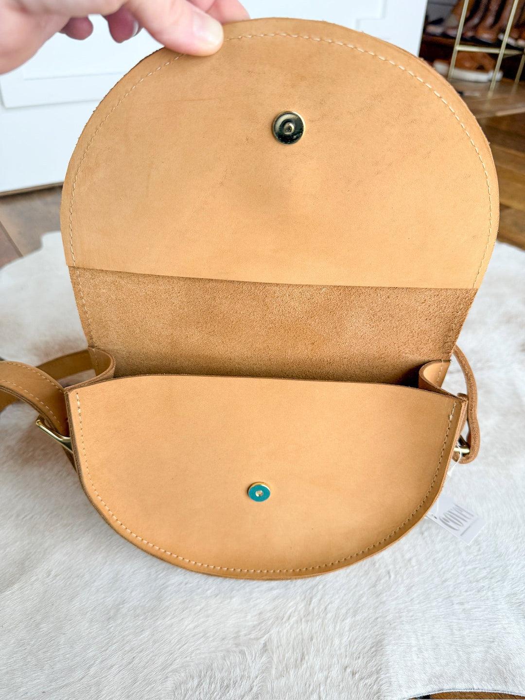 A hand holds open The Texola by Savannah Sevens western life{&}style, showing its suede interior and snap button. Made from full-grain saddle tan leather, the crossbody with a round flap sits on a light-colored surface.