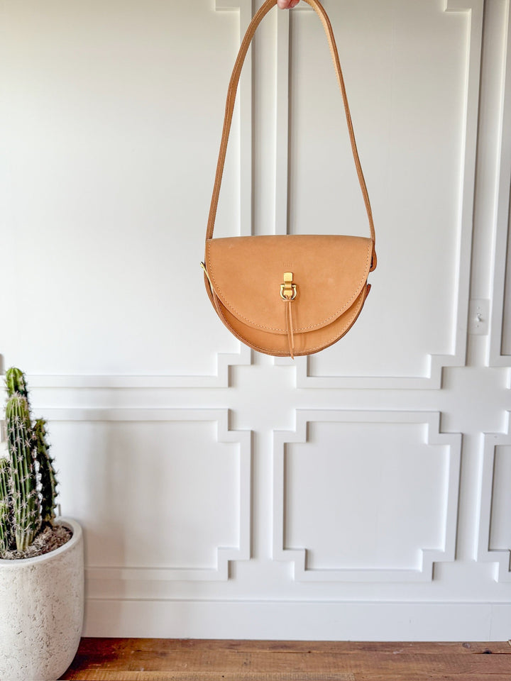The Texola by Savannah Sevens western life{&}style, a saddle tan leather crossbody with flap and gold clasp, hangs on a white paneled wall. A potted cactus rests on the wooden floor in the lower left corner.