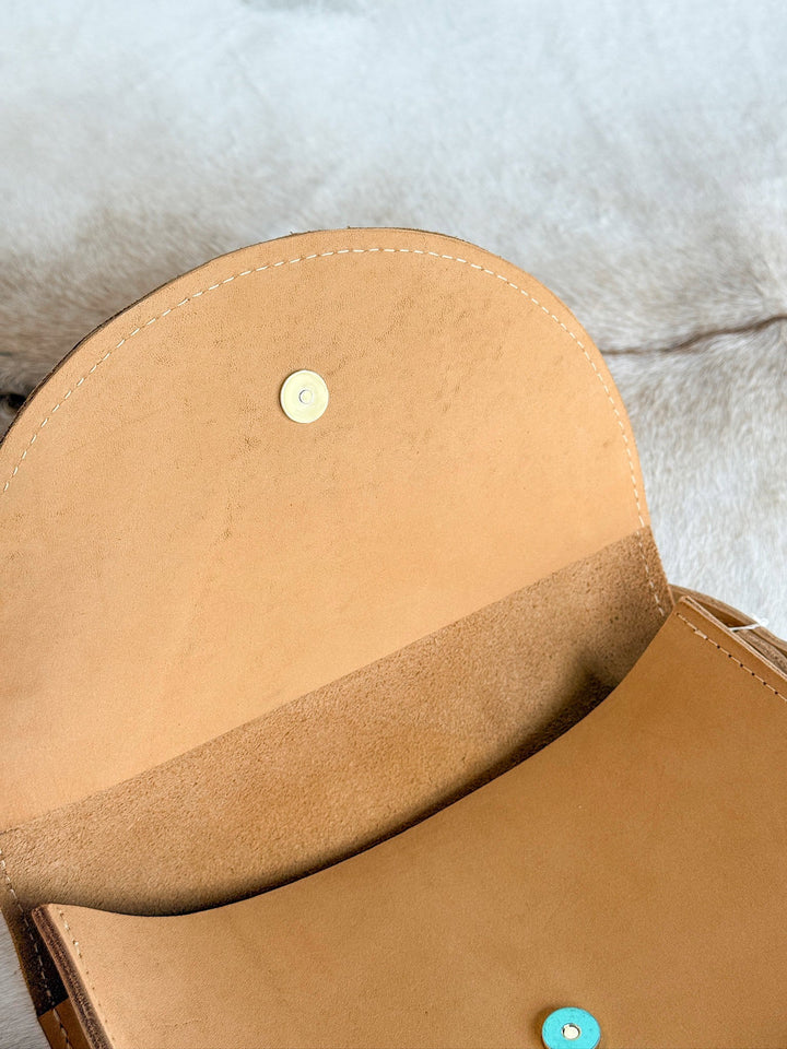 The Texola by Savannah Sevens western life{&}style is an open saddle tan bag, crafted in California from full-grain leather with a snap button closure and suede interior, shown resting on a light surface.