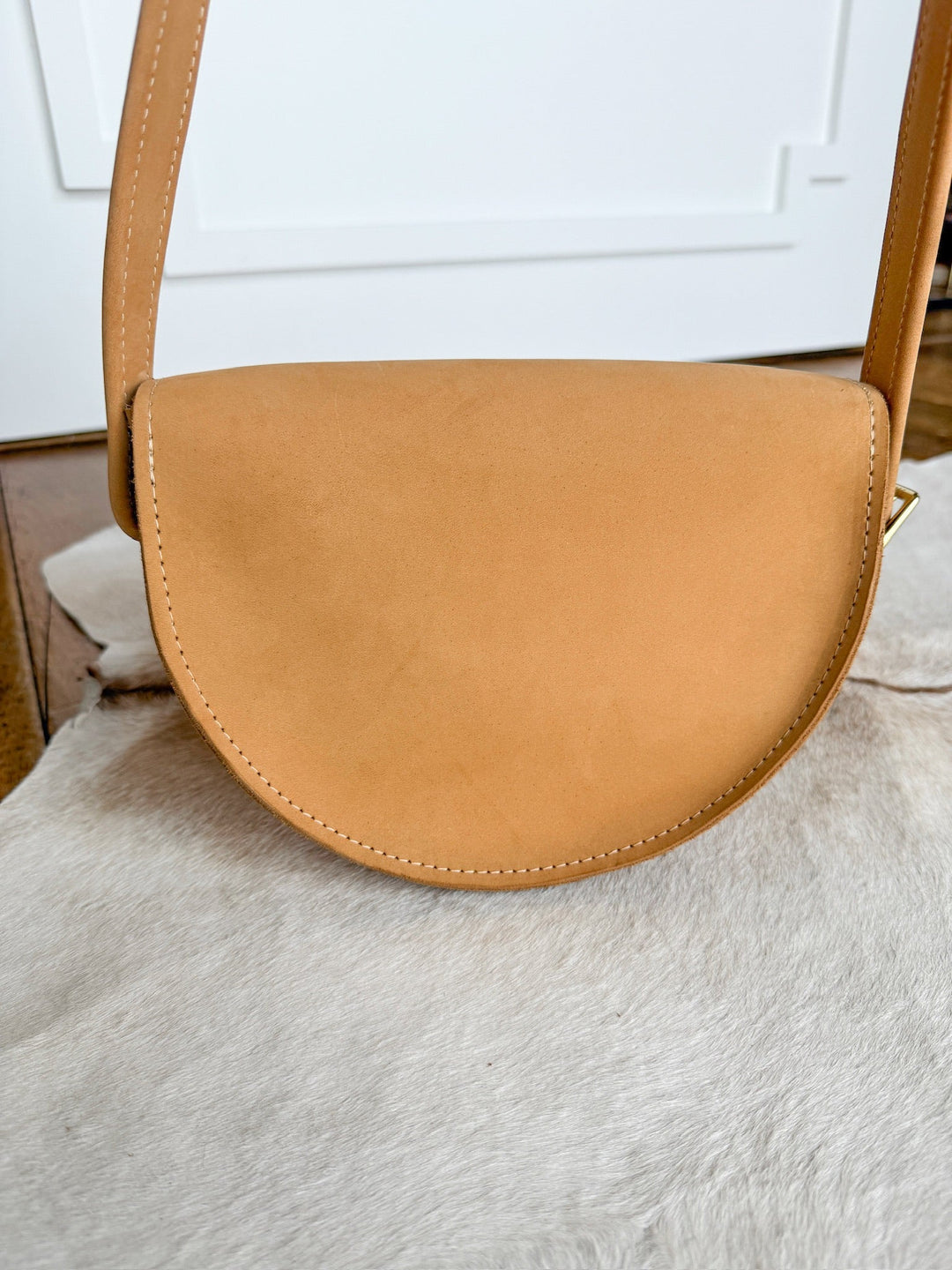 The Texola by Savannah Sevens western life{&}style is a saddle tan, full-grain leather half-moon crossbody with a wide strap, shown on a light surface against a white wall and wooden floor. Made in California. By Covet. 