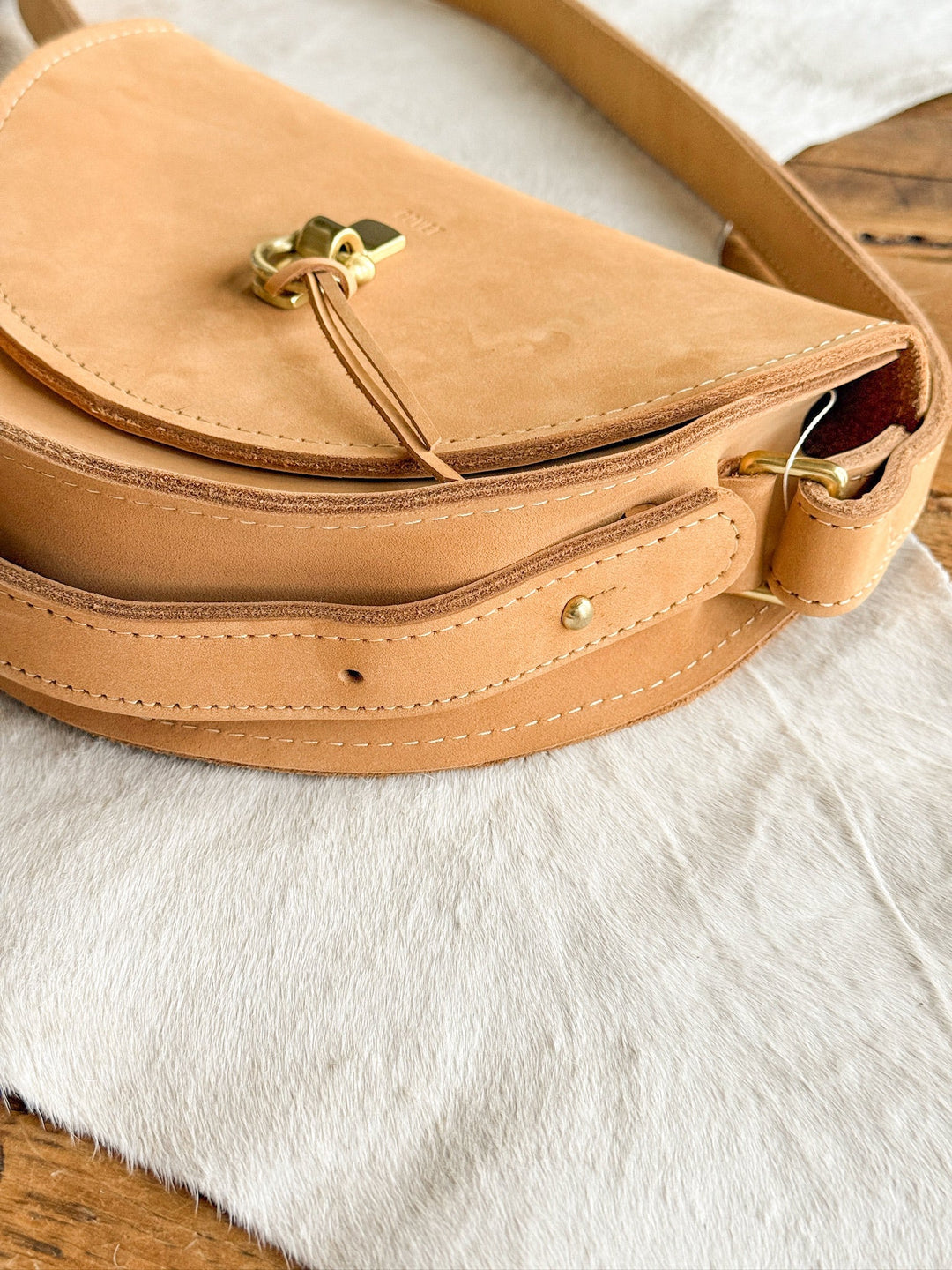 The Texola by Savannah Sevens western life{&}style is a saddle tan full-grain leather shoulder bag with gold hardware, neat stitching, and a decorative turn-lock clasp, shown on white fur and a wooden table.