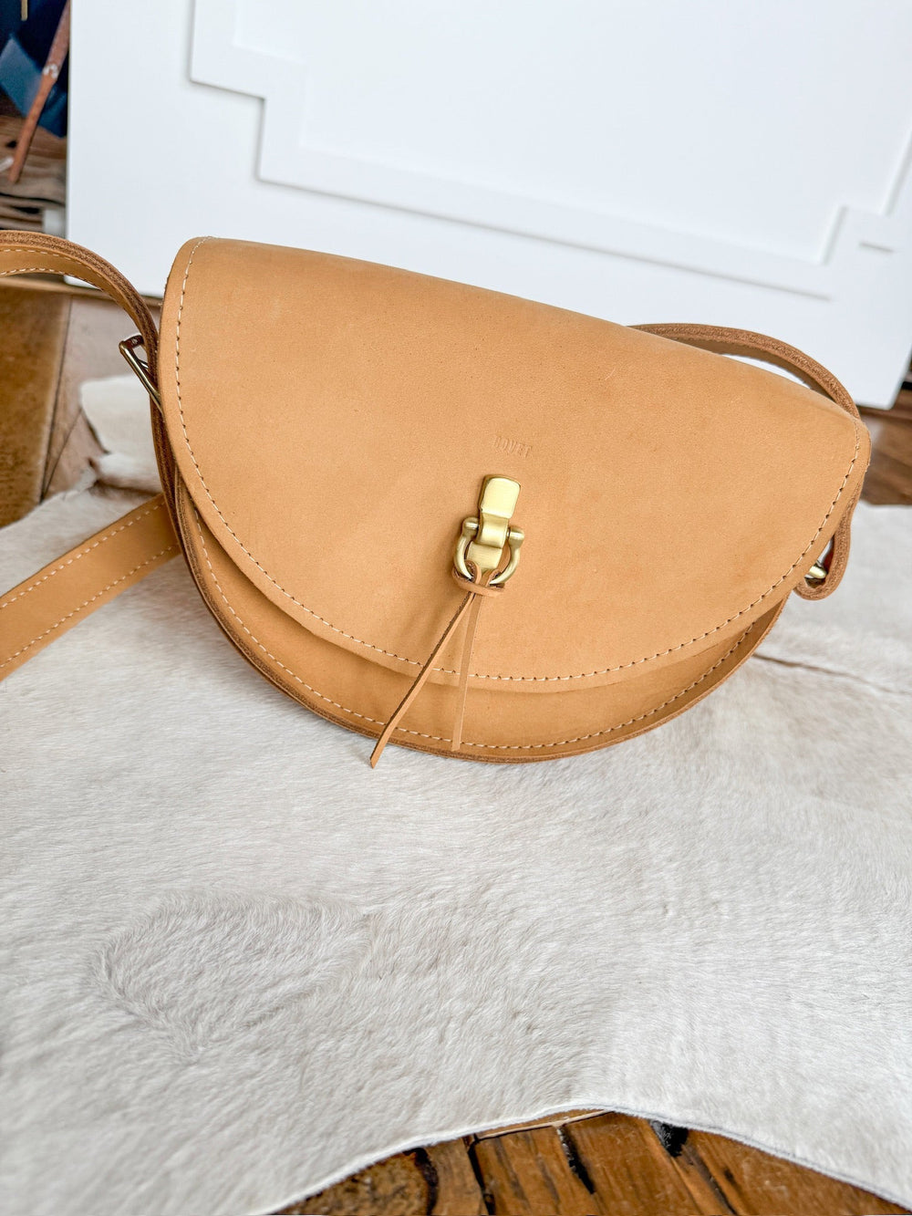 The Texola by Savannah Sevens western life{&}style, a saddle tan full-grain leather bag with rounded flap and gold clasp, rests on a light cowhide rug against a white wall.