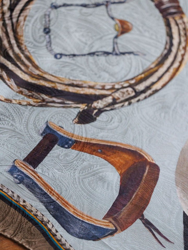 Close-up of the Texola Wild Rag by Savannah Sevens western life{&}style, a jacquard silk scarf featuring detailed coiled rope, bit, and wooden saddle illustrations on a paisley pattern inspired by classic western wild rags.