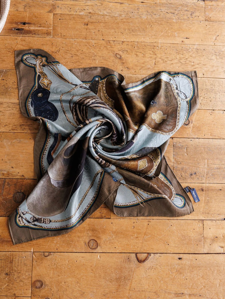 The Texola Wild Rag by Savannah Sevens western life{&}style, a jacquard silk scarf in brown, blue, and gold paisley, is artfully twisted on a rustic wooden floor with its fabric tag visible on the edge.