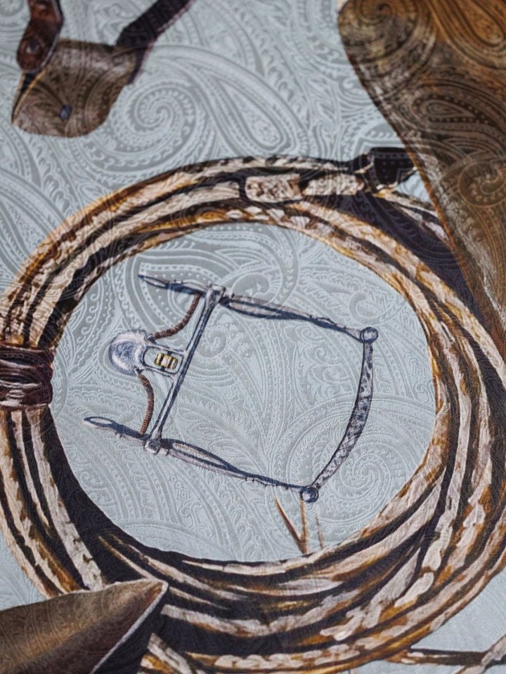A close-up of a bit and leather straps resting on the Savannah Sevens western life{&}style Texola Wild Rag, a light jacquard silk scarf with a paisley pattern that captures an authentic Western vibe.