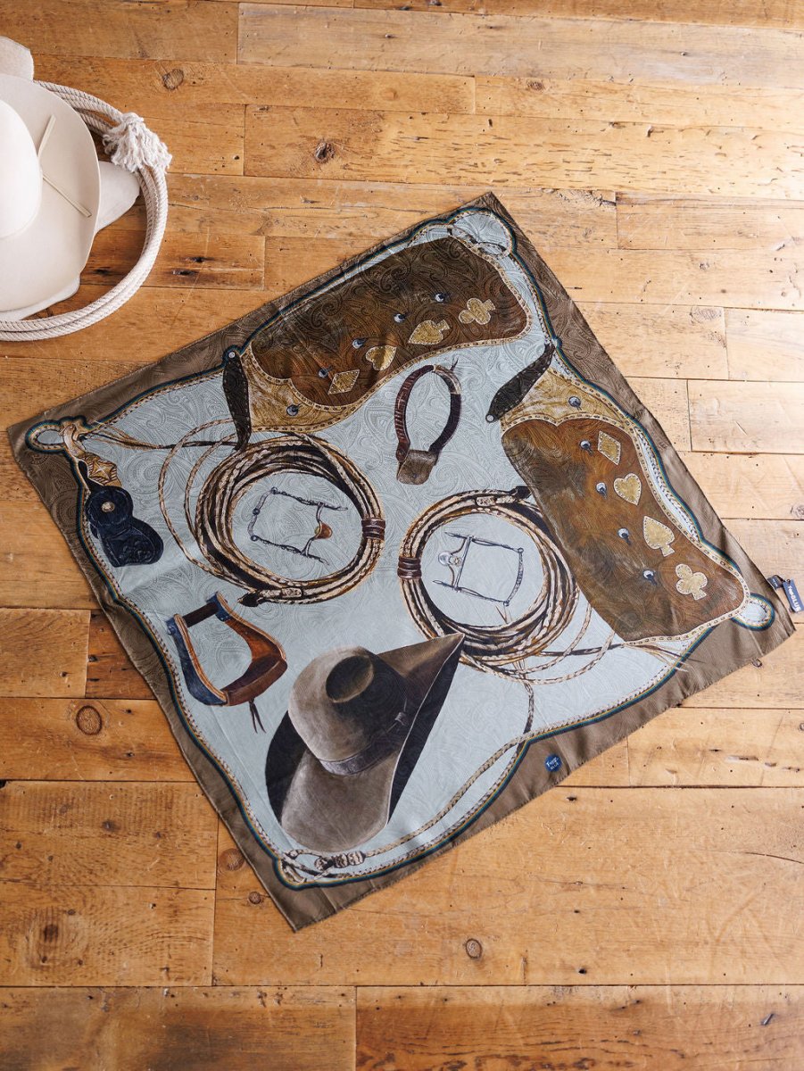 The Texola Wild Rag by Savannah Sevens western life{&}style, a square silk scarf featuring cowboy hats, boots, and saddlebags, is spread on a wooden floor next to two white hats.