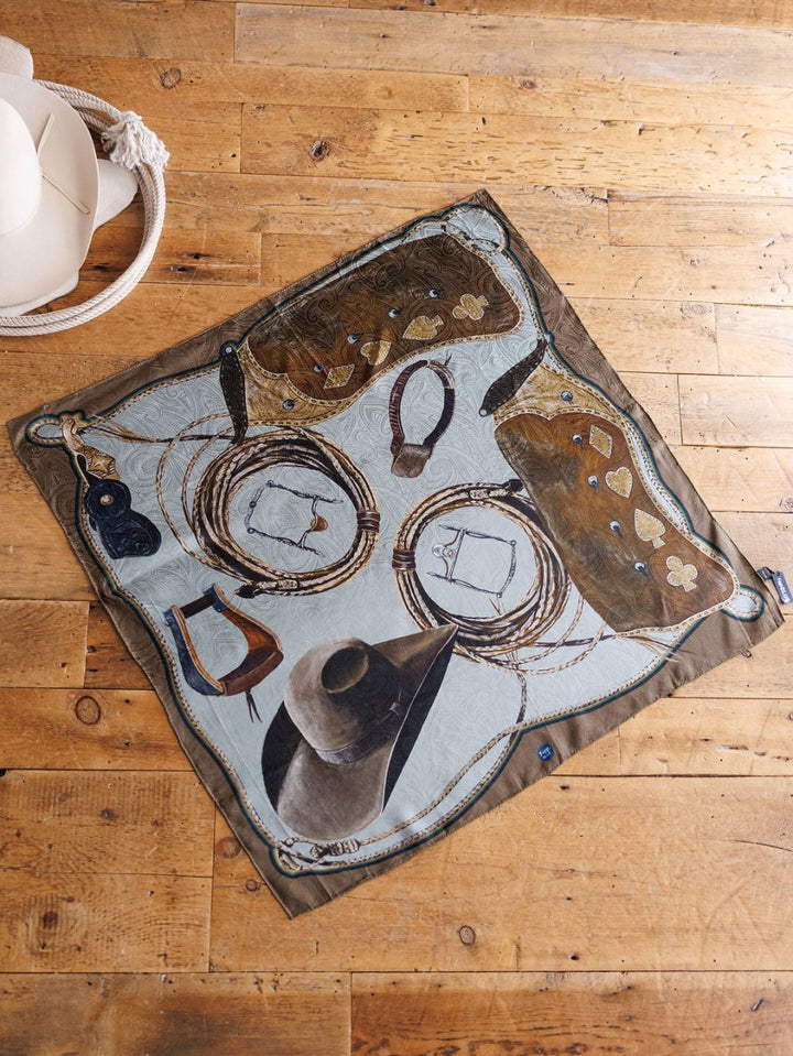 The Texola Wild Rag by Savannah Sevens western life{&}style, a square silk scarf featuring cowboy hats, boots, and saddlebags, is spread on a wooden floor next to two white hats.
