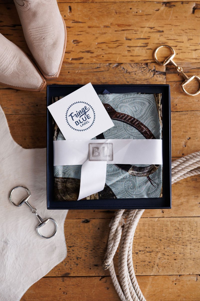 A gift box with a white ribbon and “BL” seal holds the Texola Wild Rag by Savannah Sevens western life{&}style. A “Fringe Blue” card rests on top, beside tan boots, a white fur accent, and a rope on a wooden floor.