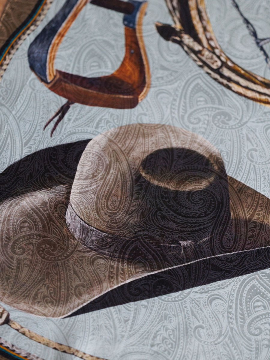 A close-up of the Texola Wild Rag by Savannah Sevens western life{&}style, featuring a brown cowboy hat and saddle on a light gray paisley jacquard silk—perfect as a western wild rag or scarf.